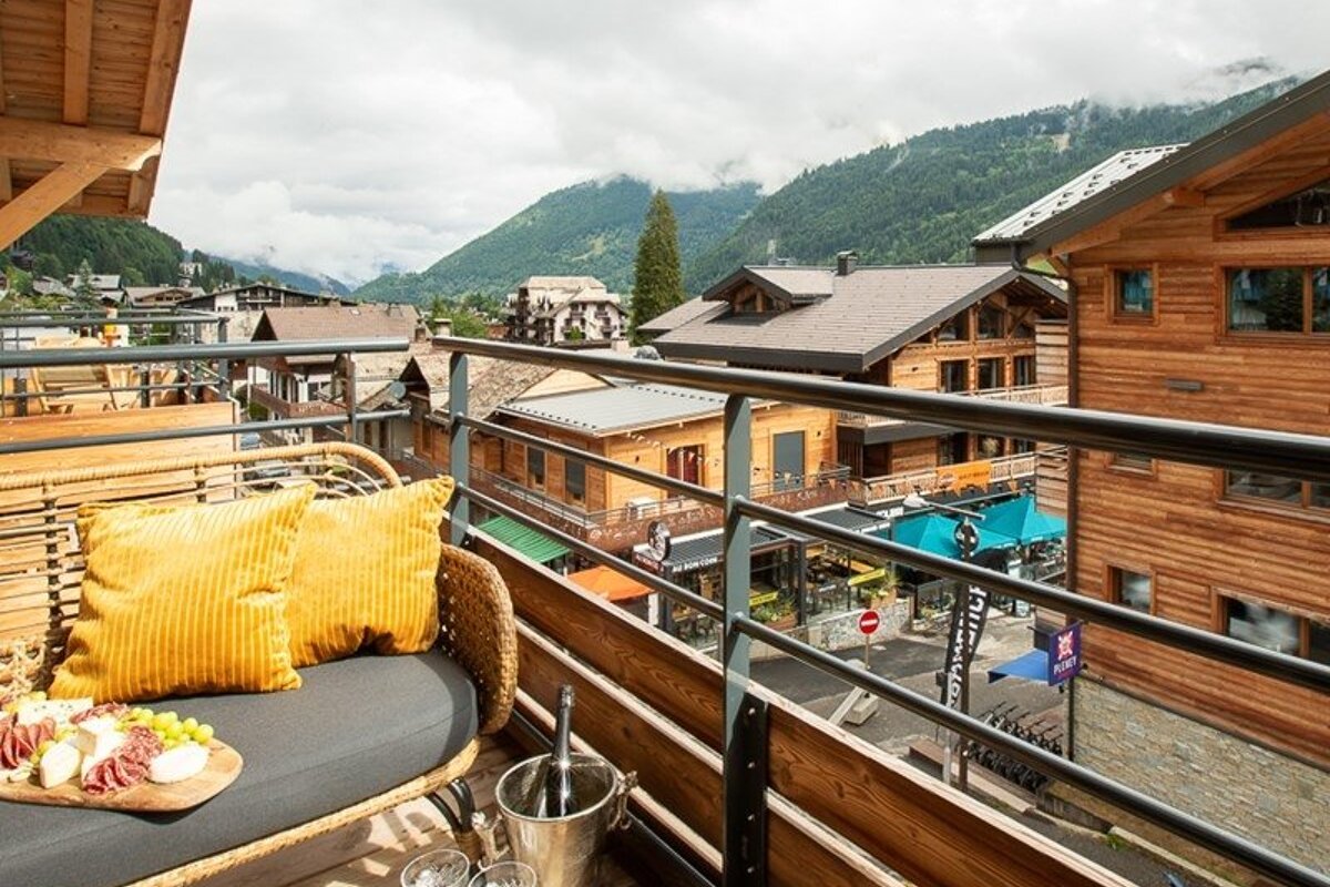 Scenic balcony with yellow cushions, snacks, and wine, overlooking a beautiful alpine town with wooden chalets and green mountains.