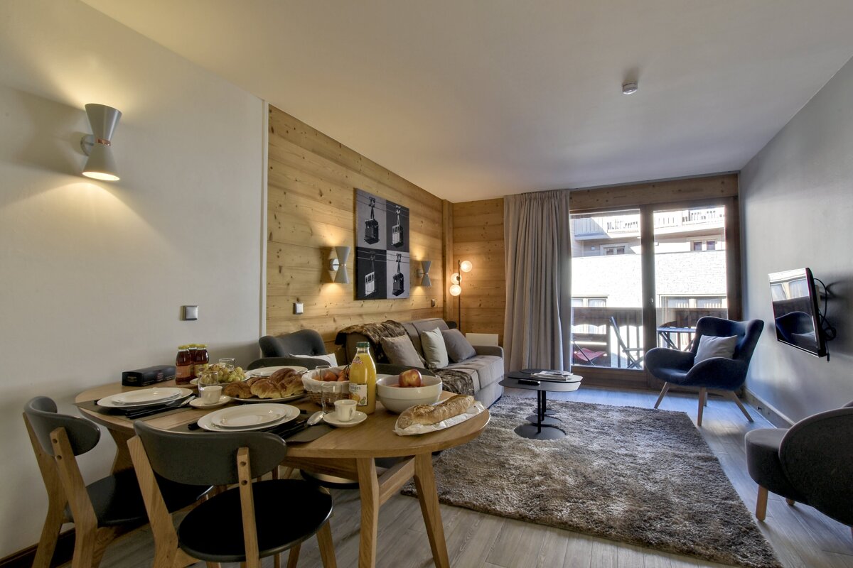 A modern, chalet-style living/dining area with a breakfast spread, wood-paneled walls, a comfortable sofa, and a balcony view.