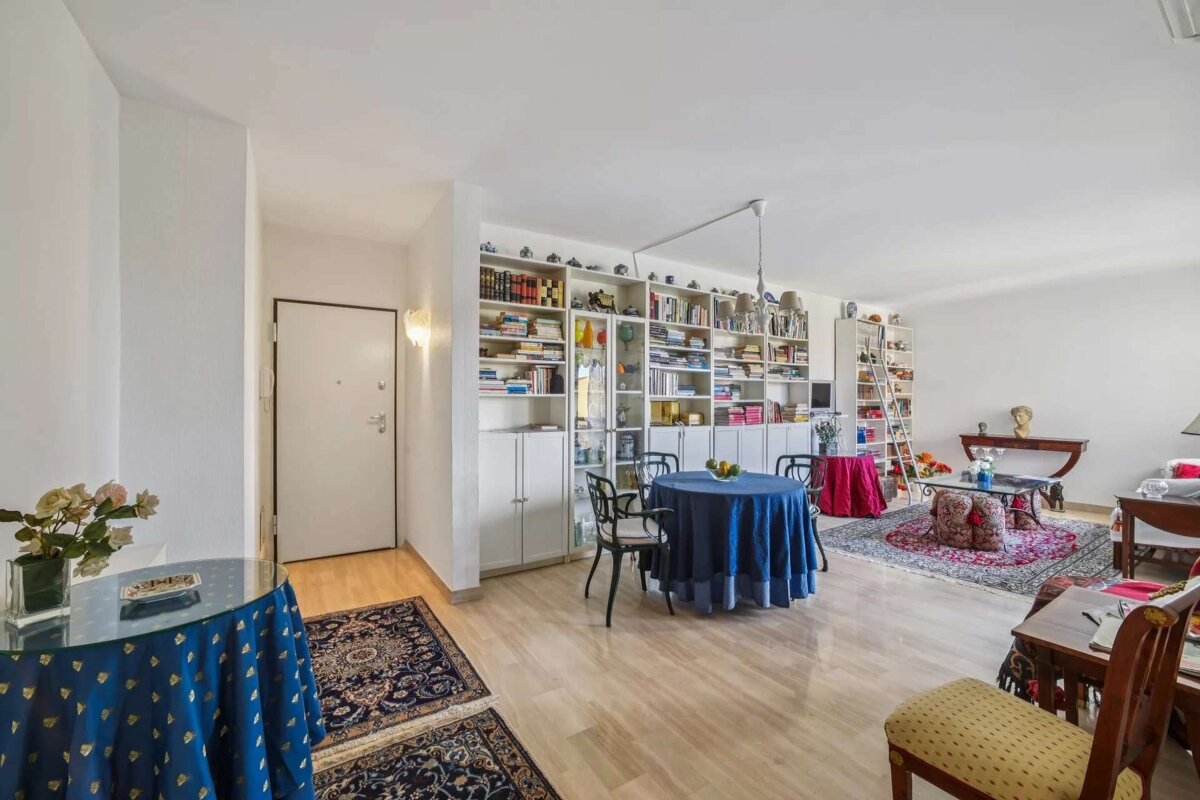 A brightly lit living room features a full wall of bookshelves, two round tables with tablecloths, light wood flooring, and ornate rugs.