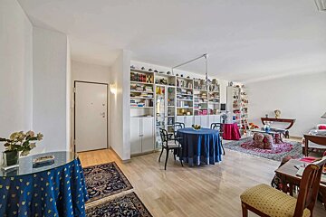 A brightly lit living room features a full wall of bookshelves, two round tables with tablecloths, light wood flooring, and ornate rugs.