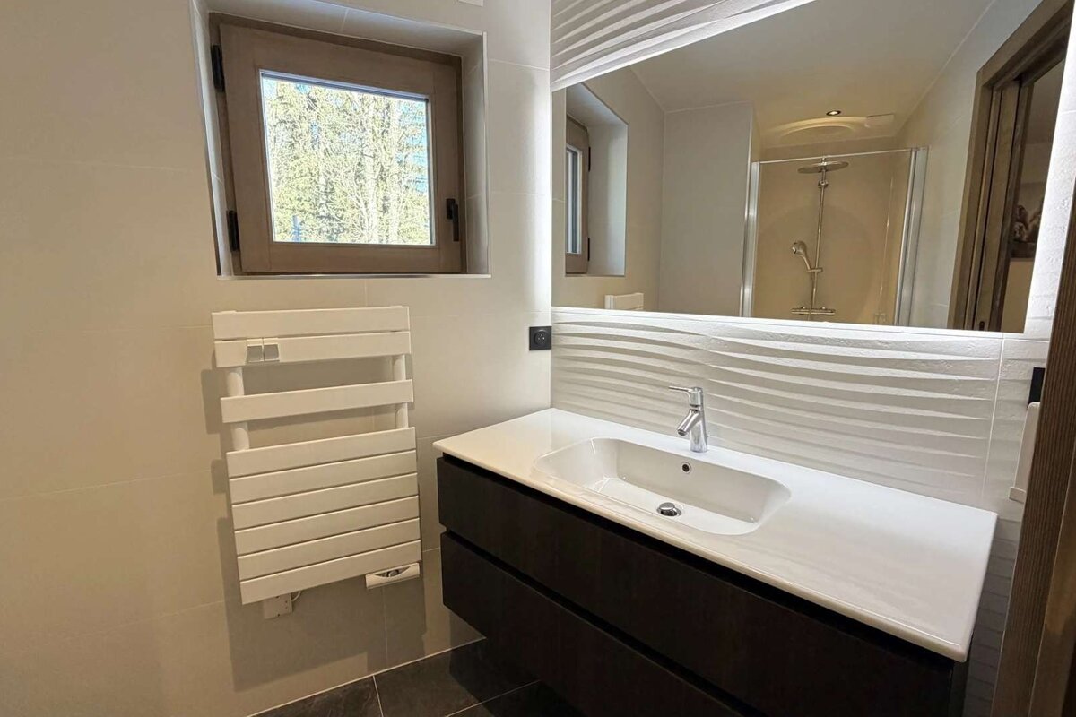 Modern bathroom with a dark vanity, white sink, large mirror, and window. Towel warmer on wall, shower visible in mirror.