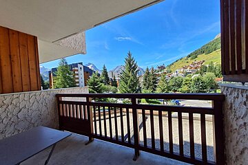 Balcony view of a sunny alpine village surrounded by green hills, pine trees, and mountains under a clear blue sky.
