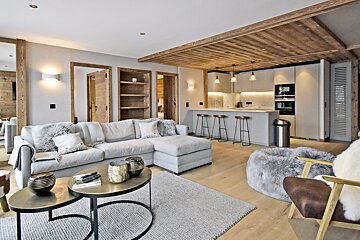 Chic modern chalet living room with a grey sectional, nested coffee tables, and a sleek kitchen under a warm wood-paneled ceiling.