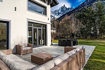 A modern house patio with outdoor seating and dining furniture, a green lawn, and towering snow-capped mountains in the background.