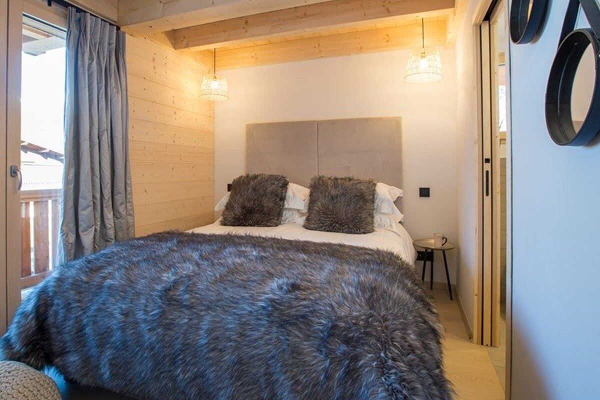 Cozy cabin bedroom featuring a bed adorned with a fluffy grey faux fur blanket, rustic wood walls, and hanging pendant lights.