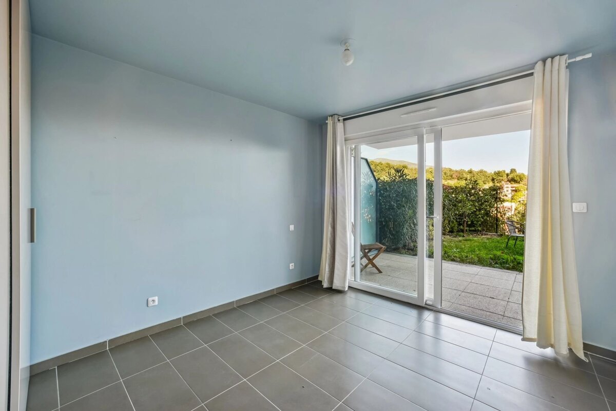 An empty room with light blue walls, gray tiled floor, and a sliding glass door opening to a patio, green garden, and distant hills.