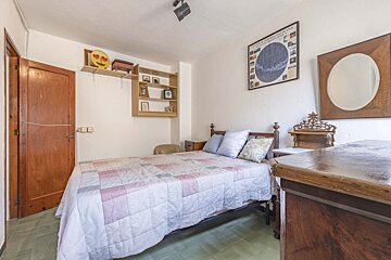 A bedroom with a bed, quilted comforter, wooden furniture, white walls, green tiled floor, and wall decor including a map and mirror.