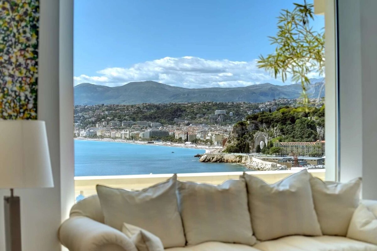 Gorgeous coastal city view from a bright room with a white sofa. Turquoise bay, urban buildings, and green mountains under a clear blue sky.