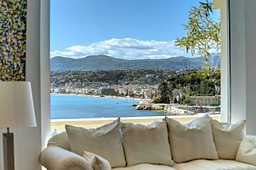 Gorgeous coastal city view from a bright room with a white sofa. Turquoise bay, urban buildings, and green mountains under a clear blue sky.