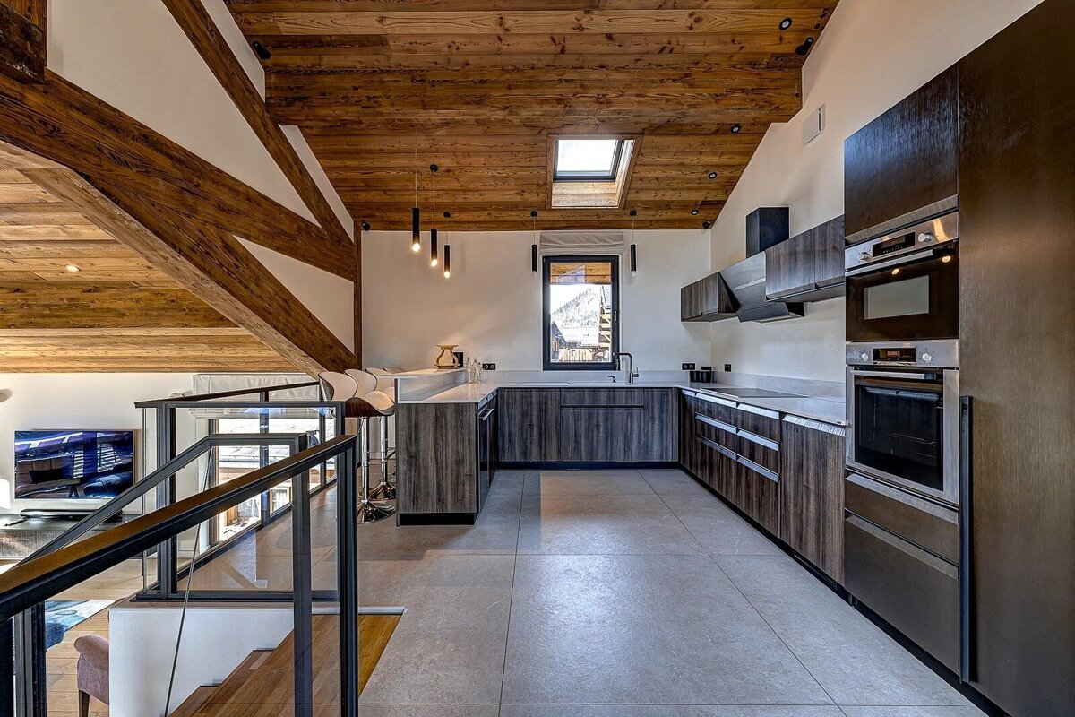 Modern loft kitchen with dark cabinets, a rustic wood-beamed ceiling with a skylight, and a glass staircase railing.