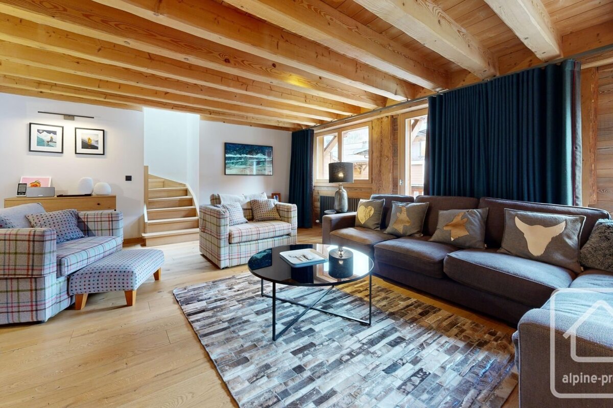 A cozy, modern chalet living room with exposed wooden beams, comfy sofas, a patterned rug, and a staircase. Features a warm, rustic design.