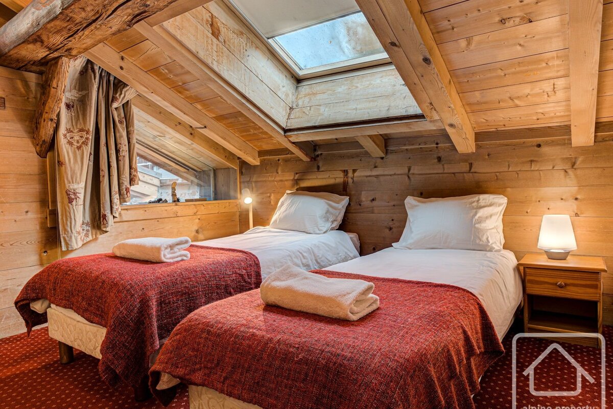 Cozy attic bedroom with two single beds, wood paneling, red blankets, and a skylight, giving a warm cabin feel.