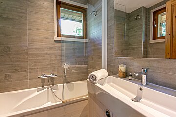 Clean, modern bathroom with a white tub and sink, grey wood-look tiles, a window, and a large mirror.