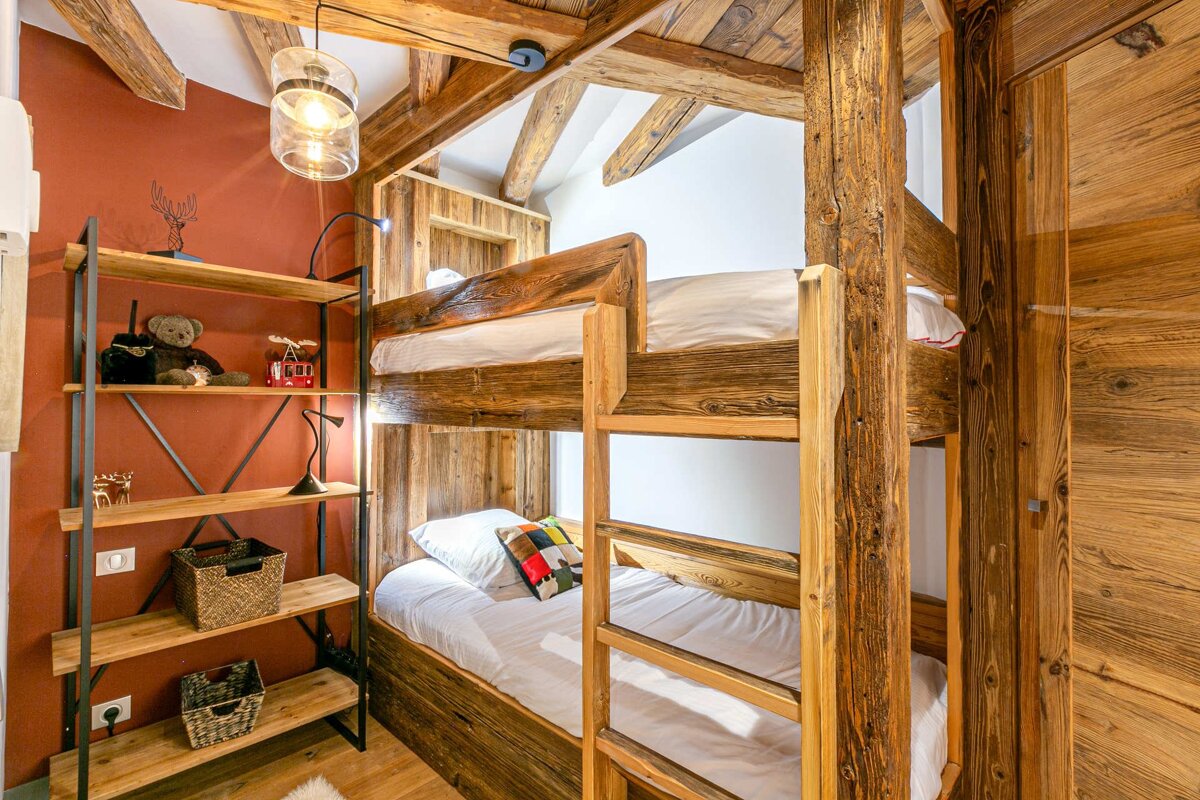 Cozy, rustic bedroom featuring sturdy wooden bunk beds, exposed ceiling beams, a warm reddish-brown wall, and a metal shelf with decor.