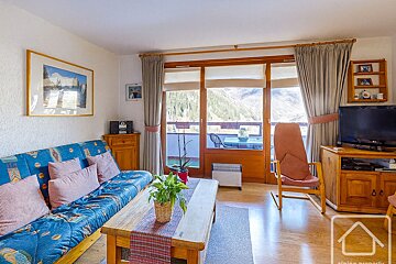 Cozy living room with a blue sofa, wooden coffee table, and large glass doors opening to a balcony with a stunning mountain view.