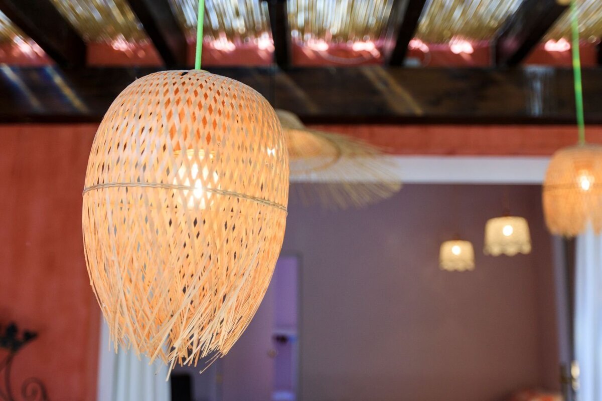 A close-up of a glowing, egg-shaped woven bamboo pendant lamp hanging from a slatted ceiling, with other blurred rustic lights in the background.