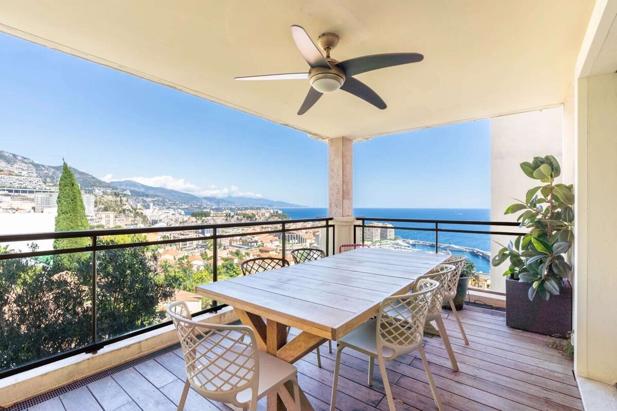 Sunny balcony with dining set & ceiling fan, overlooking a beautiful coastal city, mountains, and the sparkling blue sea.