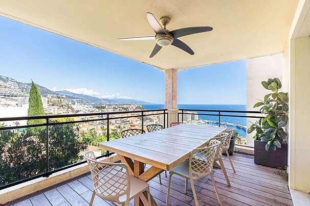 A balcony with a table, chairs, and ceiling fan, offering stunning panoramic views of a coastal city, mountains, and the blue Mediterranean Sea.