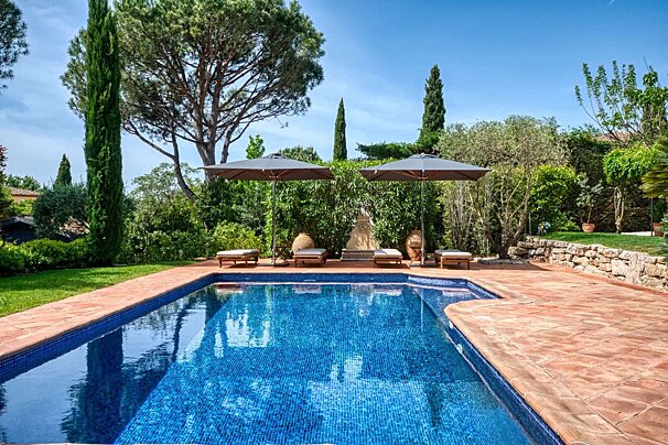 A stunning blue tiled swimming pool with terracotta surround, two umbrellas, four lounge chairs, and lush green landscaping under a bright, clear sky.
