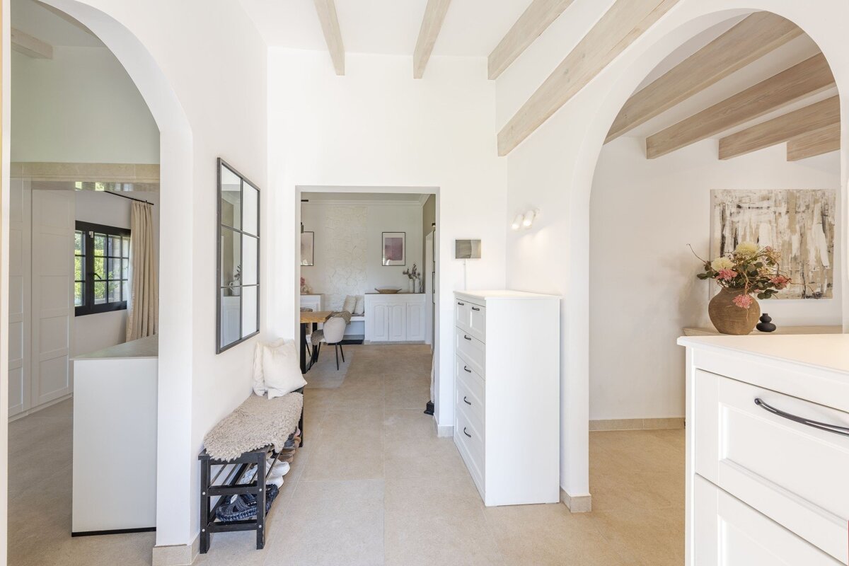Bright, arched white hallway with exposed beams, a shoe bench, storage, and an abstract painting, hinting at dining and living areas.