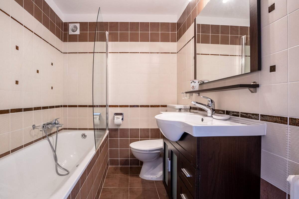 Modern bathroom with brown and beige tiles, a bathtub with glass partition, a toilet, and a sink with dark wood cabinet.