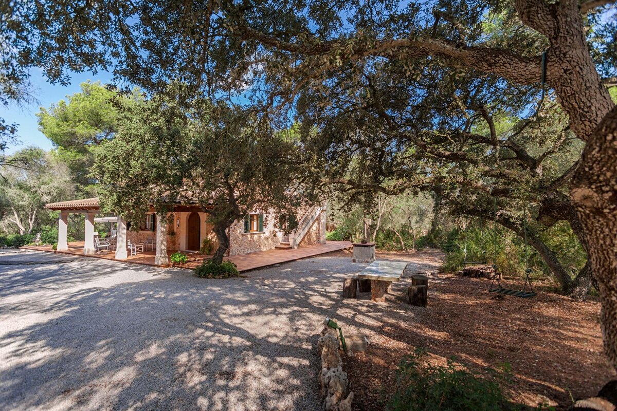 A rustic stone house with a shaded patio and gravel yard, surrounded by mature trees. Features outdoor dining and a barbecue area.
