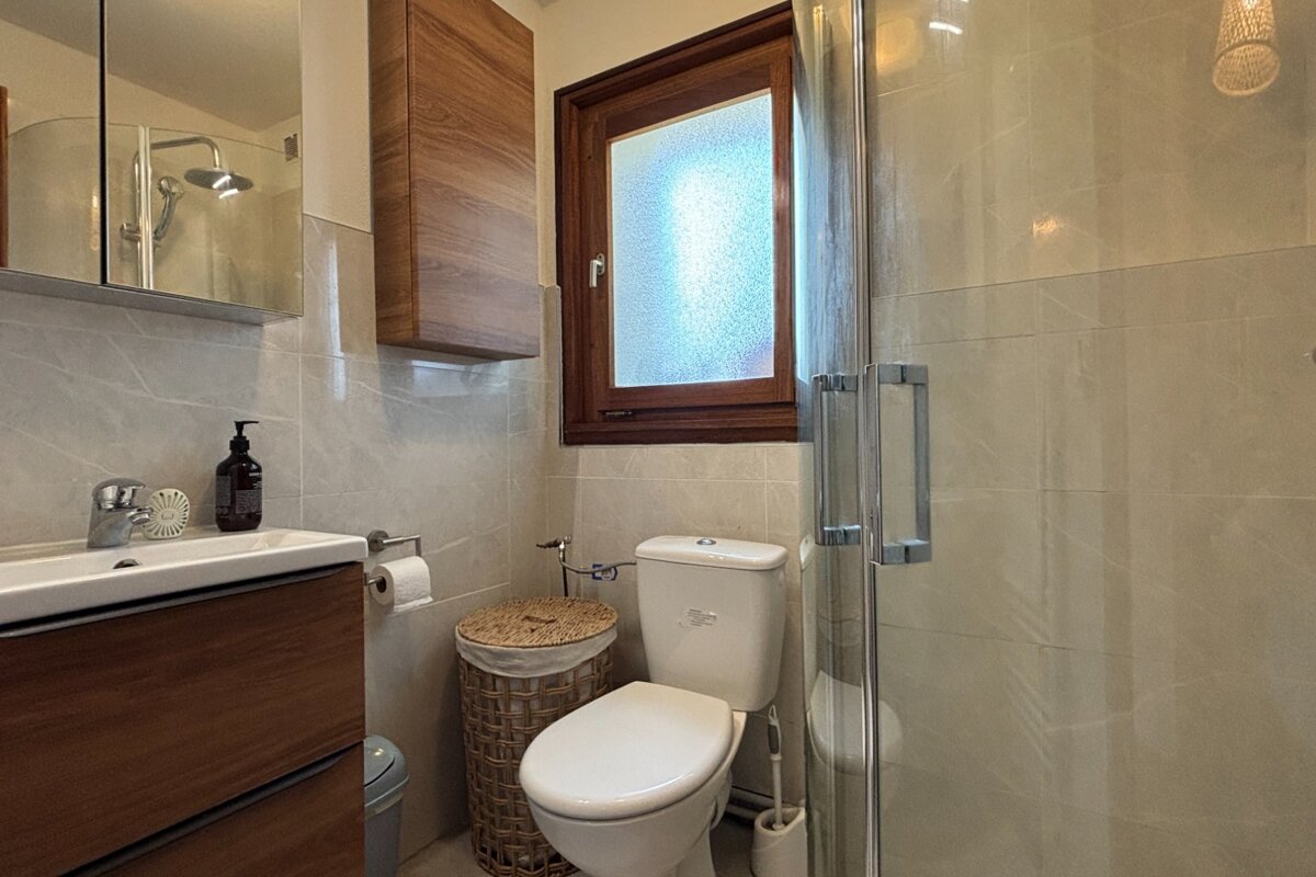 A modern bathroom featuring light tiled walls, a wooden vanity with sink, a toilet, a glass shower, and a frosted window.