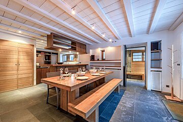 A modern rustic dining area features a long wooden table with a bench, a kitchen bar, exposed white ceiling beams, and dark stone flooring.