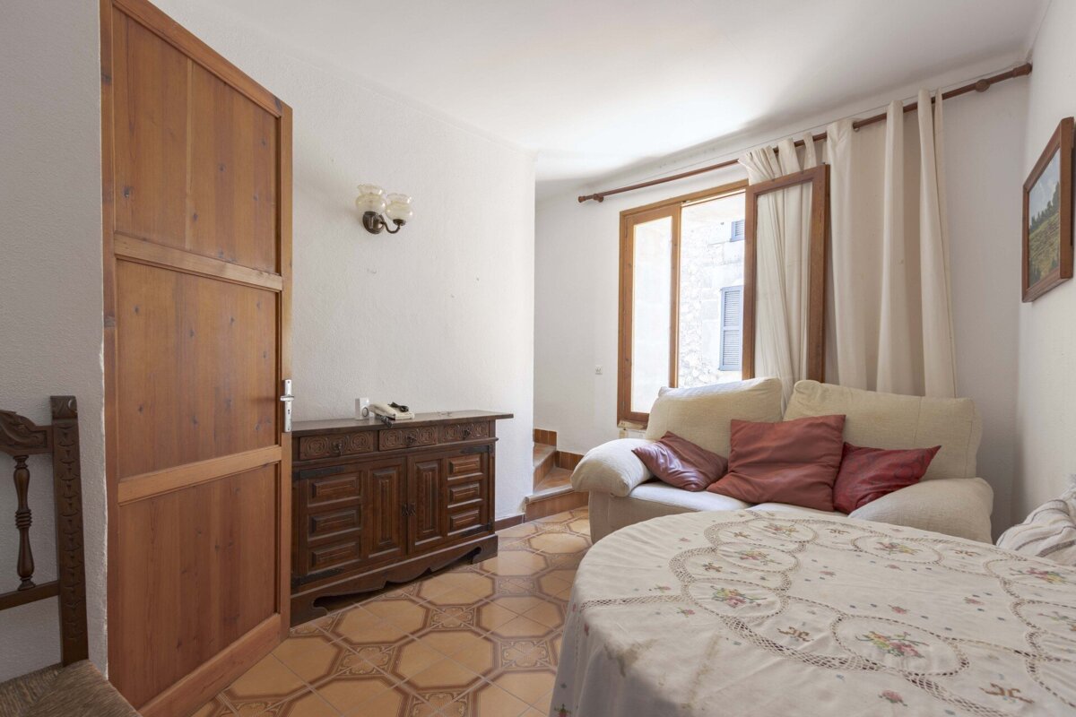 A traditional living space with a wooden door, dark cabinet, tiled floor, cream sofa, and a round, draped table by a sunlit window.