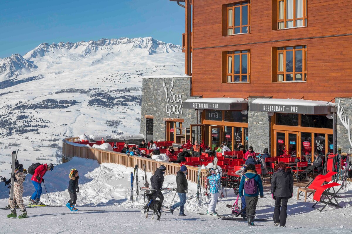 Sunny day at a bustling VOGA COGA ski resort restaurant/pub terrace overlooking vast snowy mountains, with skiers enjoying the landscape.