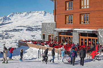 Sunny day at a bustling VOGA COGA ski resort restaurant/pub terrace overlooking vast snowy mountains, with skiers enjoying the landscape.