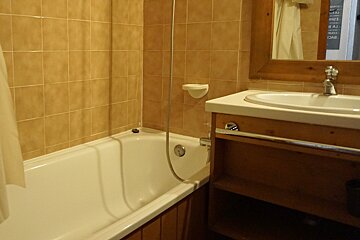 A bathroom with tan tiled walls, a white bathtub, and a shower. A wooden vanity with a sink and mirror is on the right.