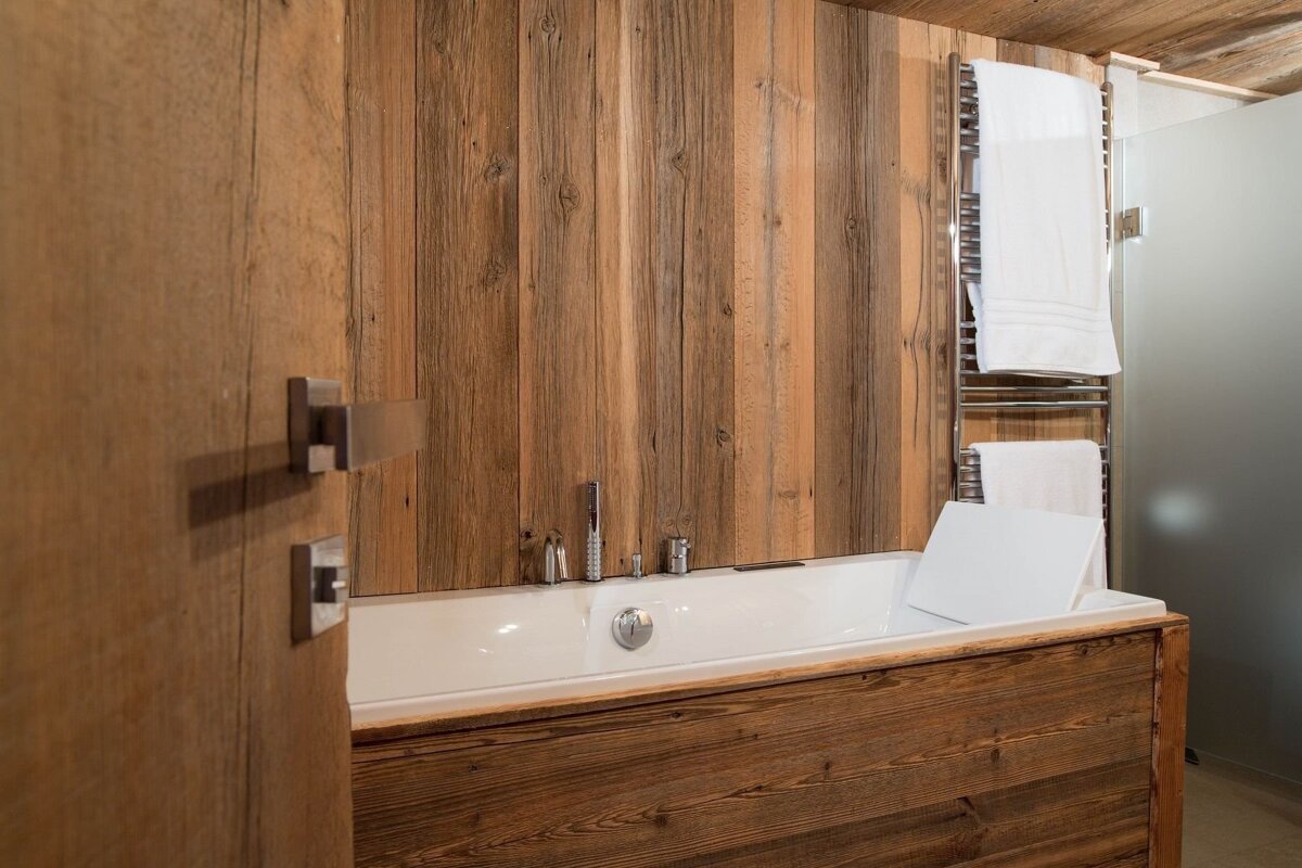 A modern bathroom features a white built-in bathtub, extensive warm wooden paneling, chrome fixtures, and a towel warmer.