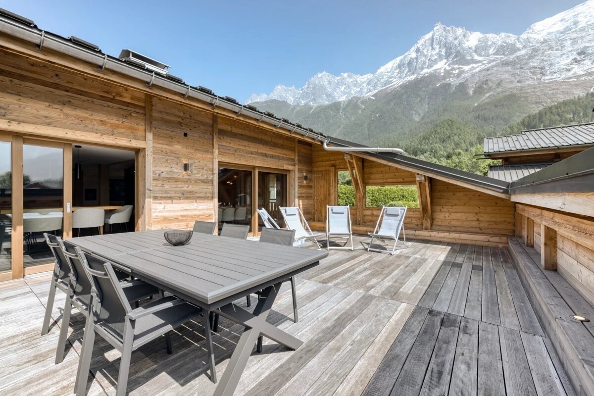 A spacious wooden deck with a dining set and lounge chairs, offering a breathtaking view of snow-capped mountains.