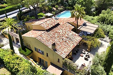 Aerial shot of a large yellow Mediterranean-style villa with red tile roofs, multiple patios, a swimming pool, and lush green gardens.