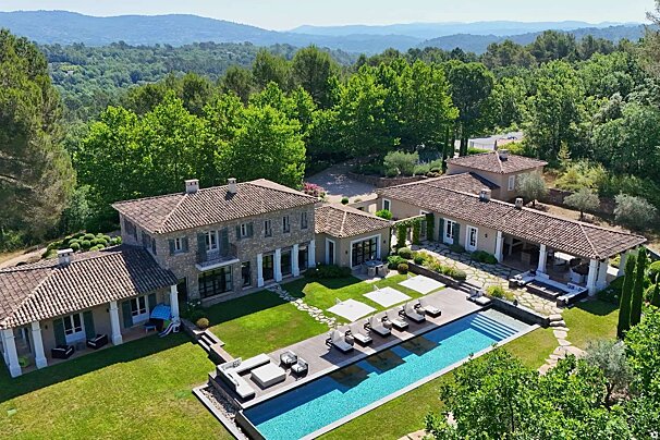 A grand Provençal-style estate with several buildings, a large swimming pool, and expansive green lawns, set against a backdrop of lush forests and distant mountains.