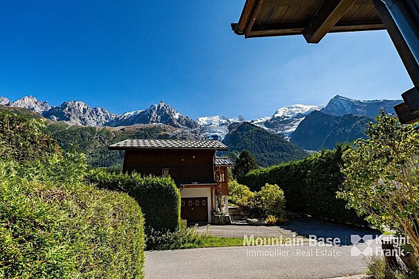A wooden house nestled among green hedges, with towering snowy mountains and a bright blue sky.