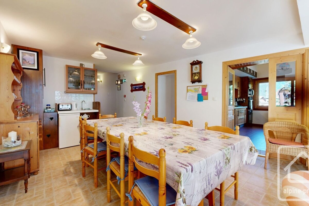 Rustic dining room with a long wooden table, floral tablecloth, matching chairs, tiled floor, and kitchen entrance.