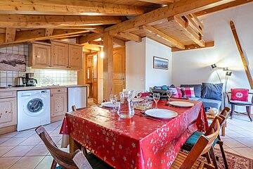 A warm, rustic chalet interior with exposed wooden beams, an open kitchen, a dining table with a red tablecloth, and a comfortable living area.