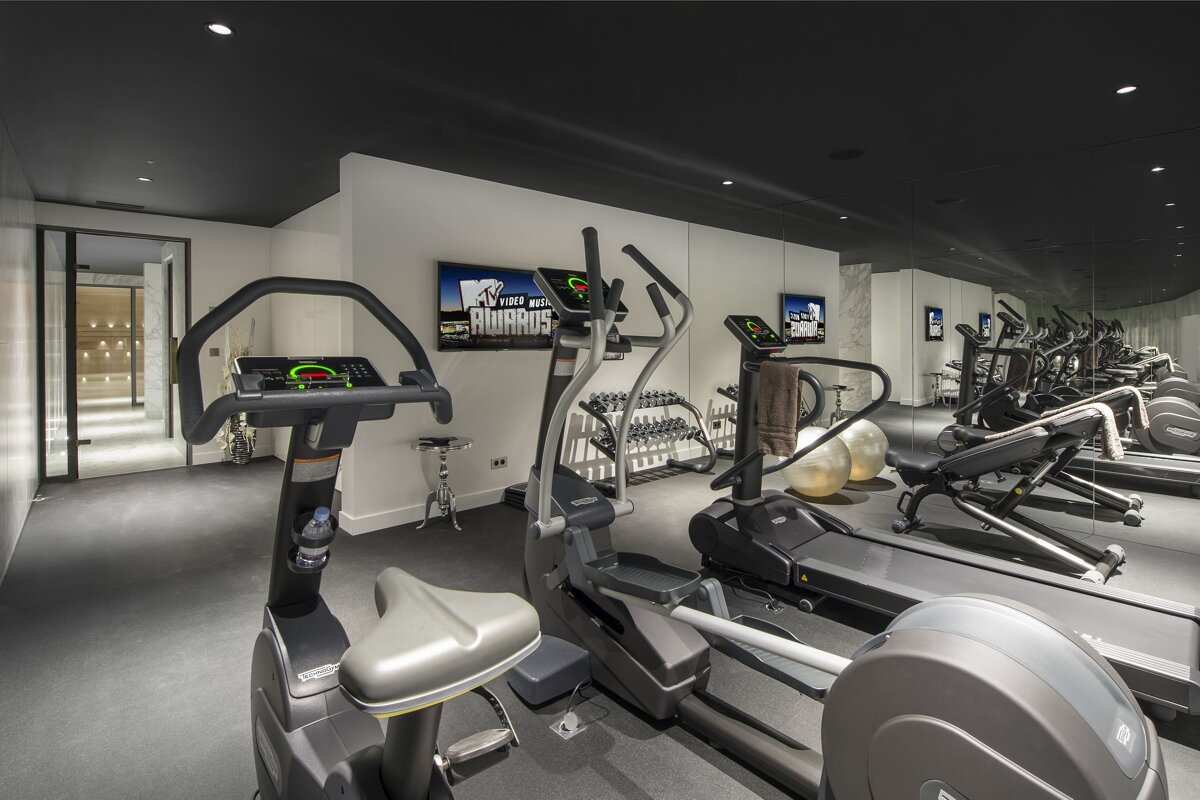A modern home gym with cardio machines like a stationary bike, elliptical, and treadmill, dumbbells, exercise balls, and TVs playing MTV.