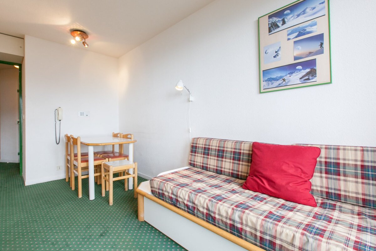 A room with green carpet, white walls, a plaid daybed with red pillow, a small dining table, and ski pictures on the wall.