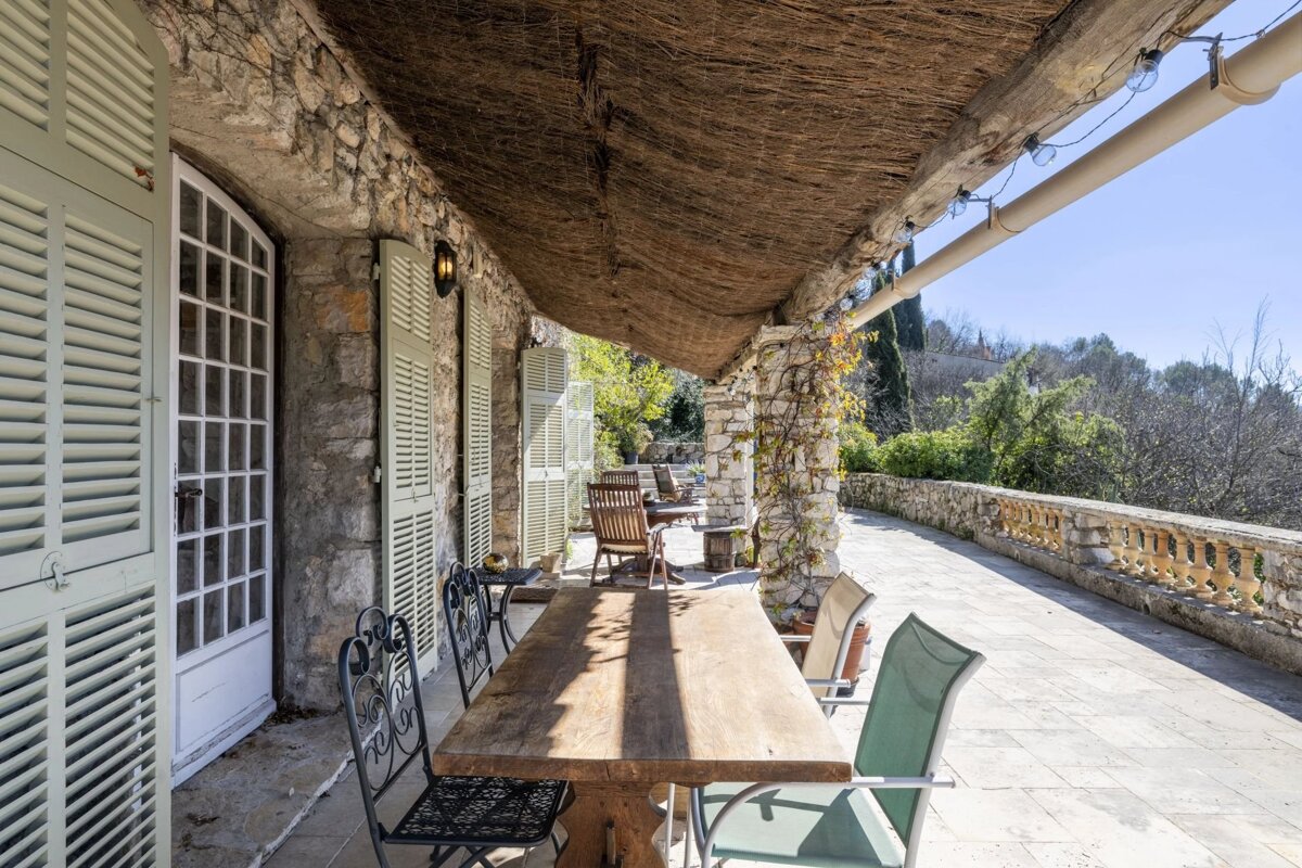 Sunny rustic stone patio with a long wooden dining table, light green shutters on a stone building, and a natural canopy.