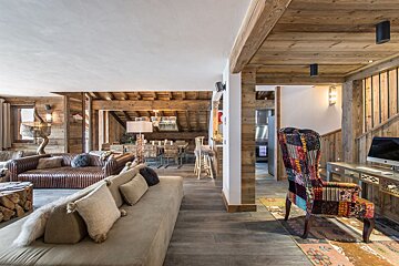 A cozy, wood-filled chalet interior featuring an open-plan living room with plush sofas, a dining area, a bar, and a unique patchwork armchair.