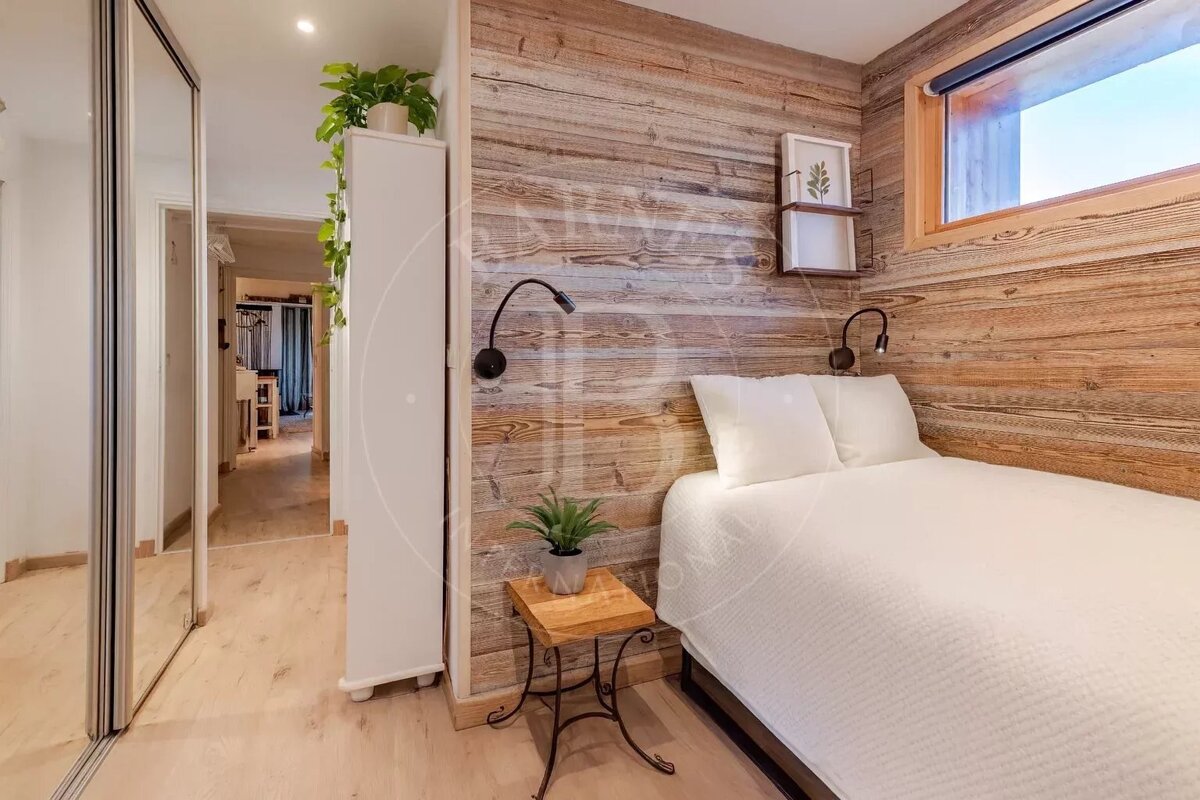 A cozy bedroom features a bed against a rustic wood-paneled wall with a window, a plant on a side table, and a hallway reflected in a mirrored closet.