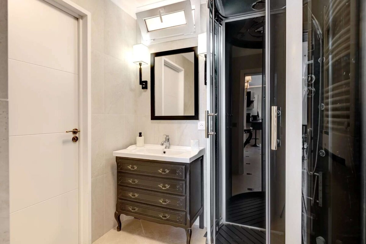 A bathroom with a white door, antique vanity, sink, mirror, and a dark glass shower stall.