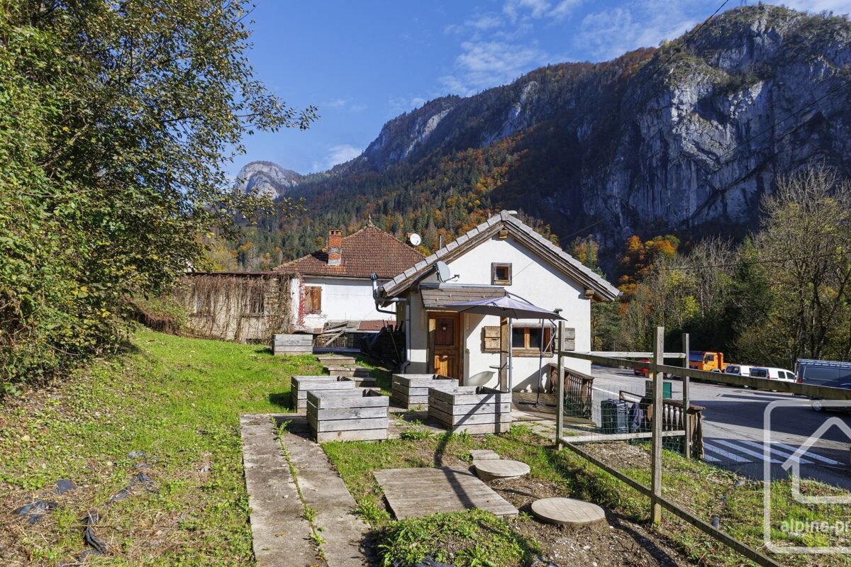 Two houses sit in a sunny alpine valley, backed by steep, rugged mountains with vibrant autumn foliage under a clear blue sky.