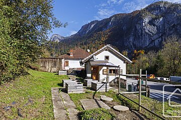 Two houses sit in a sunny alpine valley, backed by steep, rugged mountains with vibrant autumn foliage under a clear blue sky.
