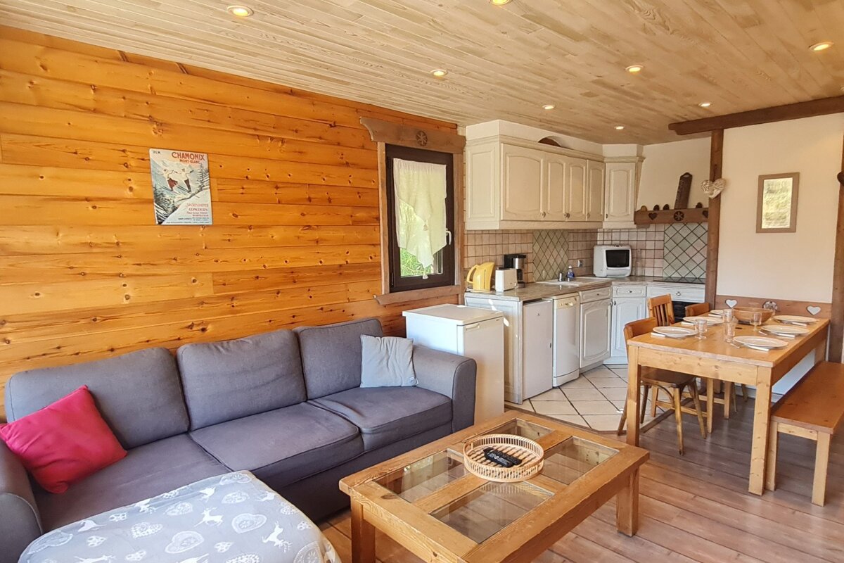 An open-plan living space with wood-paneled walls and ceiling. It features a grey sectional sofa, wooden coffee table, white kitchen, and a dining table set.