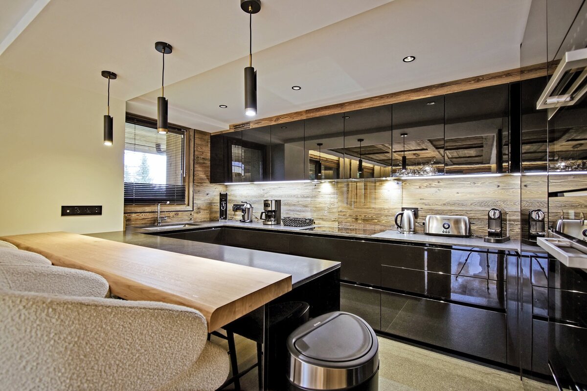 A chic modern kitchen featuring glossy black cabinets, rustic wooden walls, a light wood breakfast bar, and stylish pendant lighting. Stainless appliances accent the space.