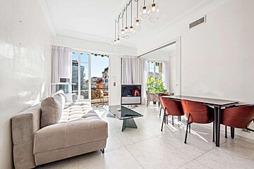 Bright, modern living room with a light sofa, glass coffee table, and dining area. Large glass doors open to a balcony with a city view.